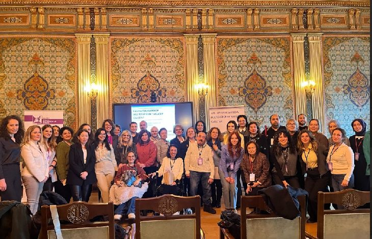 Görselde, tarihi ve gösterişli bir salonda düzenlenen bir çalıştay sonrası çekilmiş büyük bir grup fotoğrafı yer alıyor. Katılımcılar, ön planda tekerlekli sandalyedeki bireyler de dahil olmak üzere farklı yaş ve profillerden oluşuyor. Arka planda bir ekran ve yanında KASDER (Kas Hastalıkları Derneği) ile ENUYGAR logolarının yer aldığı afişler bulunuyor. Ekranda “Kas Hastalıklarında Çok Disiplinli Merkez Modelleri Çalıştayı” başlığı dikkat çekiyor.
