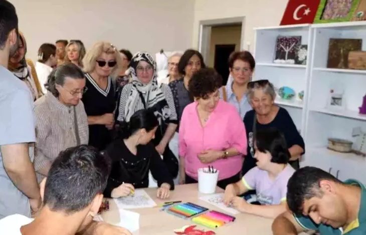 Görselde, bir masanın etrafında oturan engelli bireyler kağıt üzerine resim yapıyor. Engelli bireylerin çevresini ise yaşça büyük çoğunluğu kadın olan bir grup olduğu görülüyor.