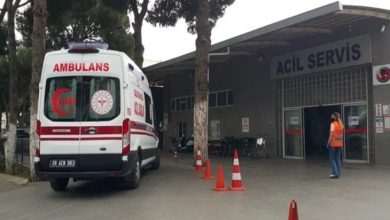 Görselde, bir hastanenin acil servis kapısı görülmektedir. Kapının önünde bir ambulans ve görevli bir erkek yer almaktadır.