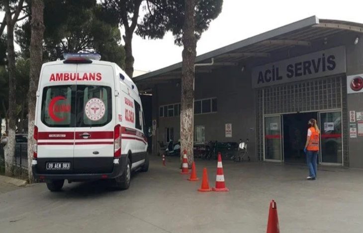 Görselde, bir hastanenin acil servis kapısı görülmektedir. Kapının önünde bir ambulans ve görevli bir erkek yer almaktadır.