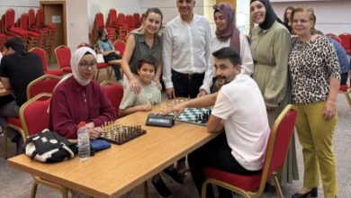 Görselde, bir satranç turnuvası sırasında çekilmiş sıcak ve samimi bir an yer alıyor. Önde bir kadın ve bir erkek satranç oynarken, etraflarında çocuklar ve yetişkinlerden oluşan bir grup onları izliyor. Oturan kadının başörtülü olduğu, gözlük taktığı ve önünde su şişesi ile çantası olduğu görülüyor. Ayakta duran izleyiciler gülümseyerek poz veriyor. Arka planda ise boş sandalyeler dikkat çekiyor.