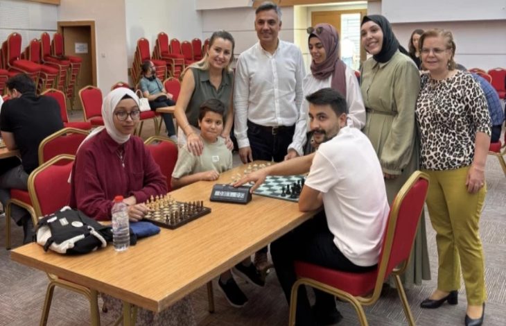 Görselde, bir satranç turnuvası sırasında çekilmiş sıcak ve samimi bir an yer alıyor. Önde bir kadın ve bir erkek satranç oynarken, etraflarında çocuklar ve yetişkinlerden oluşan bir grup onları izliyor. Oturan kadının başörtülü olduğu, gözlük taktığı ve önünde su şişesi ile çantası olduğu görülüyor. Ayakta duran izleyiciler gülümseyerek poz veriyor. Arka planda ise boş sandalyeler dikkat çekiyor.
