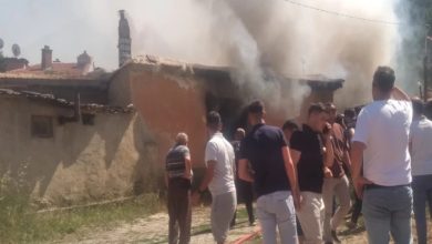 Görselde, bir evin içerisinden göğe doğru yükselen beyaz renkli duman görünüyor. Evin etrafında yangını söndürmek için toplanmış kalabalık bir grup insan yer alıyor.