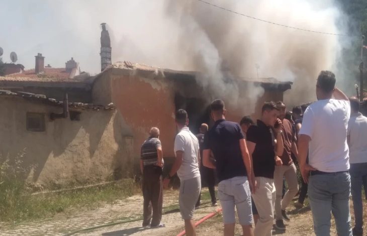 Görselde, bir evin içerisinden göğe doğru yükselen beyaz renkli duman görünüyor. Evin etrafında yangını söndürmek için toplanmış kalabalık bir grup insan yer alıyor.