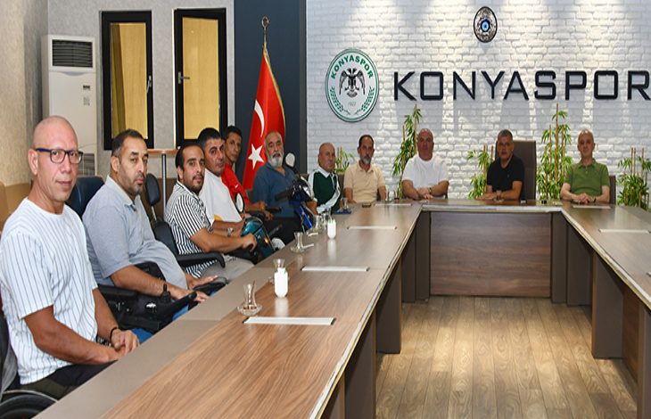 Görselde, Konyaspor logosunun yer aldığı bir toplantı salonunda çekilmiş bir kare yer alıyor. Masanın etrafında çoğu tekerlekli sandalyede oturan yaklaşık 13 kişi bulunuyor. Sol tarafta Türk bayrağı görülüyor. Duvarın arka kısmında "KONYASPOR" yazısı büyük harflerle yer almakta. Katılımcılar ciddi ve dikkatli bir şekilde kameraya poz veriyor.