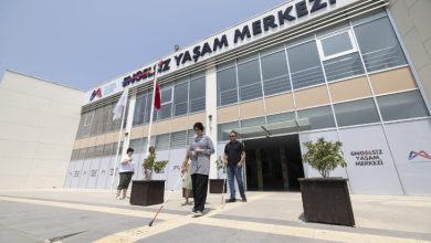 Görselde, Mersin Büyükşehir Belediyesi'ne ait "Engelsiz Yaşam Merkezi" binasının ön cephesi görülüyor. Binanın girişinde birkaç kişi yer alıyor. Ön planda, elinde beyaz baston olan bir görme engelli birey sarı takip çizgileri üzerinde yürürken dikkat çekiyor. Arkasında ona eşlik eden birkaç kişi bulunuyor.