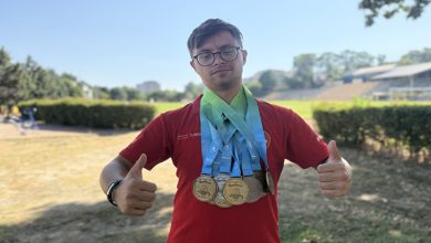 Down sendromlu milli atlet Emirhan Akçakoca’nın üstünde kırmızı tişört giyili, boynunda kazandığı madalyaları bulunuyor ve iki elinin baş parmağını kaldırarak başarı pozu veriyor.