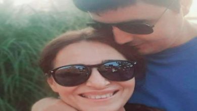 Bu fotoğrafta, yakın bir pozda çekilmiş iki kişi görülüyor. Kadın önde, gülümseyerek kameraya bakıyor; yüzünde sıcak ve içten bir ifade var. Erkek ise arkada, başını kadının başına yaslamış, onu sevgiyle sarmalamış şekilde duruyor. İkisi de güneş gözlüğü takıyor. Arka planda yeşillikler ve gün batımı ışığı var gibi görünüyor.