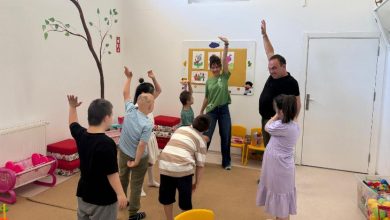 Görselde, bir sınıf ortamında düzenlenen drama atölyesinden neşeli bir an görülüyor. Çocuklar, iki yetişkin eğitmenin rehberliğinde ellerini havaya kaldırarak grup halinde bir etkinlik yapıyor. Çocukların bir kısmının Down sendromlu olduğu fark ediliyor. Duvarlar renkli çocuk çizimleriyle süslenmiş, yerde oyuncaklar ve minik sandalyeler yer almakta. Eğitmenler çocuklara enerjiyle eşlik ediyor.