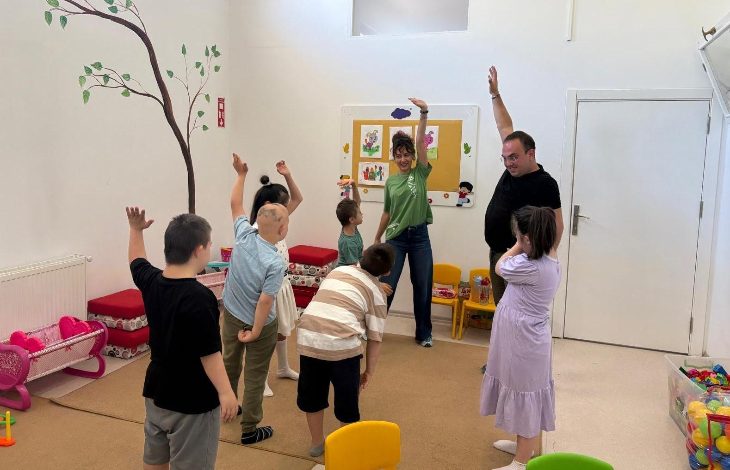 Görselde, bir sınıf ortamında düzenlenen drama atölyesinden neşeli bir an görülüyor. Çocuklar, iki yetişkin eğitmenin rehberliğinde ellerini havaya kaldırarak grup halinde bir etkinlik yapıyor. Çocukların bir kısmının Down sendromlu olduğu fark ediliyor. Duvarlar renkli çocuk çizimleriyle süslenmiş, yerde oyuncaklar ve minik sandalyeler yer almakta. Eğitmenler çocuklara enerjiyle eşlik ediyor.