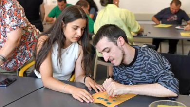 Görselde bir sınıf ortamında, özel gereksinimli bireylerle gerçekleştirilen bir sanat etkinliği görülüyor. Ön planda, genç bir kadın, tekerlekli sandalyedeki bir erkekle birlikte çalışıyor. Ellerinde renkli damgalarla süslenmiş bir kâğıt var; birlikte dikkatlice desenler oluşturuyorlar. Arka planda ise benzer etkinliklere katılan başka bireyler yer almakta.