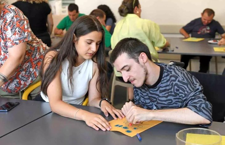 Görselde bir sınıf ortamında, özel gereksinimli bireylerle gerçekleştirilen bir sanat etkinliği görülüyor. Ön planda, genç bir kadın, tekerlekli sandalyedeki bir erkekle birlikte çalışıyor. Ellerinde renkli damgalarla süslenmiş bir kâğıt var; birlikte dikkatlice desenler oluşturuyorlar. Arka planda ise benzer etkinliklere katılan başka bireyler yer almakta.