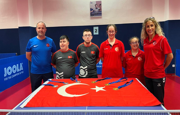 Down Masa Tenisi Erkek ve Kadın Milli Takımı sporcularımız, tenis masası üstünde bulunan Türk Bayrağı üzerine kazanılan madalyaları koyarak poz veriyor.