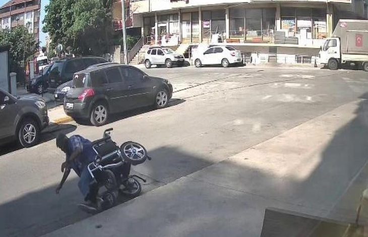Görselde, bir yokuşta akülü tekerlekli sandalyesi devrilmiş bir engelli birey (Fatih Bayrak) yere düşmüş halde görülüyor. Olay bir cadde kenarında, kaldırım hizasında gerçekleşmiş. Arka planda park etmiş araçlar ve dükkanların yer aldığı bir bina yer alıyor. Görüntü, bir güvenlik kamerasından alınmış.