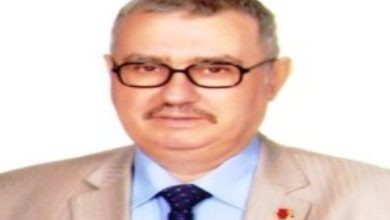 Bu görselde, orta yaşlı bir erkek beyaz arka plan önünde poz vermiş. Üzerinde açık renk bir takım elbise, mavi gömlek ve koyu renk kravat bulunuyor. Gözlük takmakta ve hafif bir gülümseme ile kameraya bakıyor. Fotoğraf, resmi veya biyografik amaçla çekilmiş bir portre izlenimi veriyor.