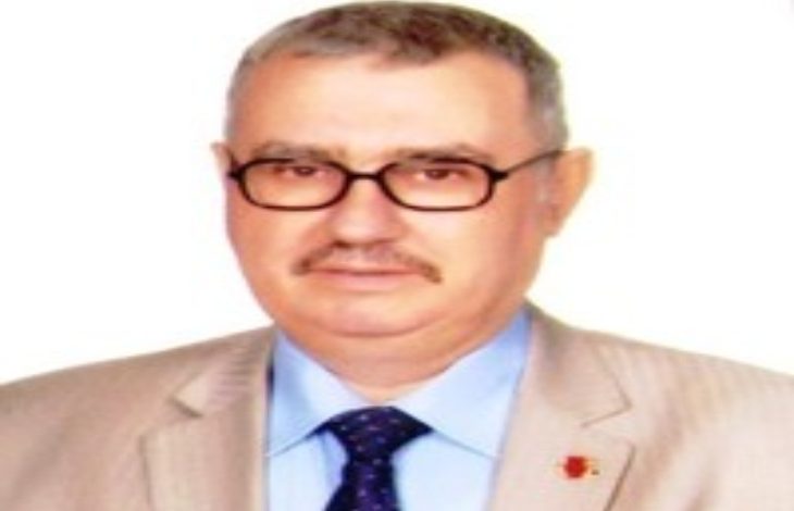 Bu görselde, orta yaşlı bir erkek beyaz arka plan önünde poz vermiş. Üzerinde açık renk bir takım elbise, mavi gömlek ve koyu renk kravat bulunuyor. Gözlük takmakta ve hafif bir gülümseme ile kameraya bakıyor. Fotoğraf, resmi veya biyografik amaçla çekilmiş bir portre izlenimi veriyor.