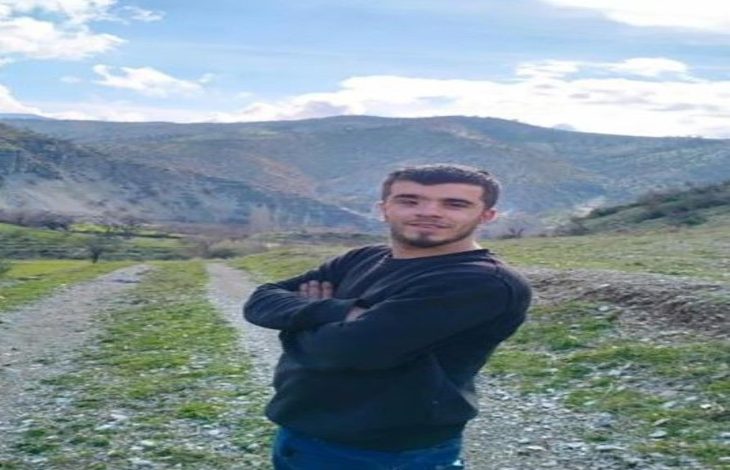 Görselde, doğa manzaralı bir kırsal alanda genç bir erkek kamera karşısında poz veriyor. Kollarını göğsünde kavuşturmuş, gülümsüyor. Üzerinde siyah bir kazak ve mavi pantolon var. Arka planda dağlar, tepeler ve açık mavi gökyüzü dikkat çekiyor. Zemin taşlık ve otlarla kaplı bir patika yolu gösteriyor.