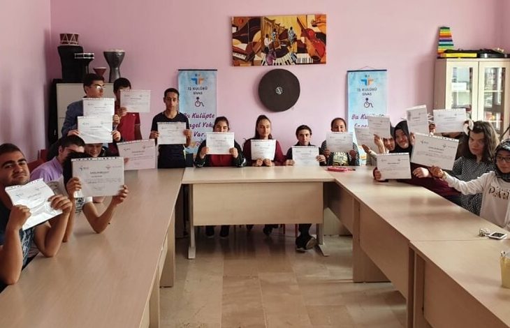 Görselde, bir sınıf ortamında "sertifika töreni" havası yansıtılıyor. U şeklinde yerleştirilmiş masaların etrafında oturan 15'e yakın genç birey, ellerinde sertifikalarını gururla tutuyor. Arkada iki afiş yer alıyor: Üzerinde “İş Kulübü” ve “Bu Kulüpte Engel Yok” ifadeleri okunabiliyor. Duvarda renkli bir müzik temalı tablo asılı, odanın dekorasyonu sade ve duvarlar pembe renkte.