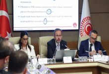 Görselde, Türkiye Büyük Millet Meclisi’nde yapılan bir komisyon toplantısı görülüyor. Masanın başında AK Parti İzmir Milletvekili Mehmet Muharrem Kasapoğlu yer alıyor ve konuşma yapıyor. Sol ve sağ yanında yer alan katılımcılar notlar alıyor veya belge inceliyor. Arkada dev bir ekran üzerinde “Engelli Sağlık Kurulu Raporları” ve “yaklaşık 2,5 milyon” gibi ifadeler okunabiliyor. Sol tarafta Türk bayrağı, sağda ise TBMM amblemi içeren bir bayrak bulunuyor.