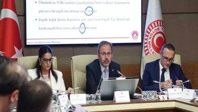 Görselde, Türkiye Büyük Millet Meclisi’nde yapılan bir komisyon toplantısı görülüyor. Masanın başında AK Parti İzmir Milletvekili Mehmet Muharrem Kasapoğlu yer alıyor ve konuşma yapıyor. Sol ve sağ yanında yer alan katılımcılar notlar alıyor veya belge inceliyor. Arkada dev bir ekran üzerinde “Engelli Sağlık Kurulu Raporları” ve “yaklaşık 2,5 milyon” gibi ifadeler okunabiliyor. Sol tarafta Türk bayrağı, sağda ise TBMM amblemi içeren bir bayrak bulunuyor.