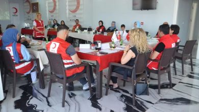 Bu fotoğraf, Türk Kızılay Muş Şubesi tarafından düzenlenen bir konferans veya eğitim toplantısını göstermektedir. Katılımcılar dikdörtgen masa düzeninde oturmuş, çoğu Türk Kızılay yeleği giymiş durumda.
