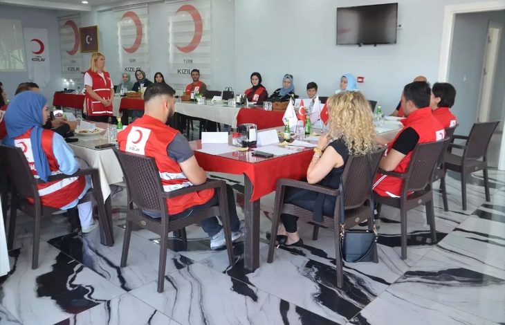 Bu fotoğraf, Türk Kızılay Muş Şubesi tarafından düzenlenen bir konferans veya eğitim toplantısını göstermektedir. Katılımcılar dikdörtgen masa düzeninde oturmuş, çoğu Türk Kızılay yeleği giymiş durumda.