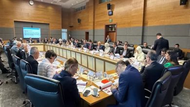 Görselde, Türkiye Büyük Millet Meclisi’nde (TBMM) gerçekleştirilen resmi bir toplantı yer alıyor. Uzun, dikdörtgen bir masanın etrafında çok sayıda milletvekili ve katılımcı oturmakta. Herkesin önünde belgeler, kitapçıklar ve isimlikler var. Bazı kişiler not alıyor, bazıları konuşmaları dinliyor. Odanın arka tarafında büyük bir ekran, duvarda saat ve dijital gösterge yer alıyor.