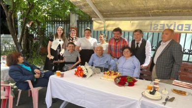 Görselde, açık havada düzenlenen bir doğum günü kutlaması görülüyor. Ortada yaşlı bir adam (Prof. Dr. Coşkun Özdemir) oturuyor; önünde çiçekler ve mumlarla süslenmiş bir doğum günü pastası var. Sağında ve solunda farklı yaş gruplarından kadın ve erkekler oturuyor ve ayakta duruyor. Masada plastik bardaklar, tabaklar ve ikramlar bulunuyor. Üstte “BELEDİYESİ” yazısının göründüğü bir tente yer almakta.