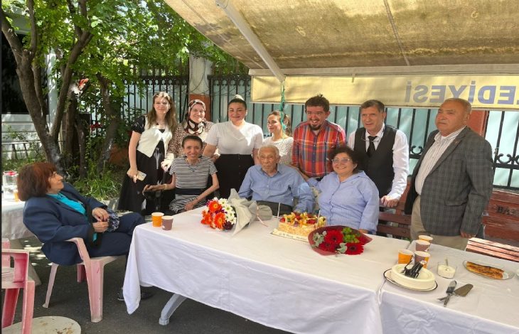 Görselde, açık havada düzenlenen bir doğum günü kutlaması görülüyor. Ortada yaşlı bir adam (Prof. Dr. Coşkun Özdemir) oturuyor; önünde çiçekler ve mumlarla süslenmiş bir doğum günü pastası var. Sağında ve solunda farklı yaş gruplarından kadın ve erkekler oturuyor ve ayakta duruyor. Masada plastik bardaklar, tabaklar ve ikramlar bulunuyor. Üstte “BELEDİYESİ” yazısının göründüğü bir tente yer almakta.