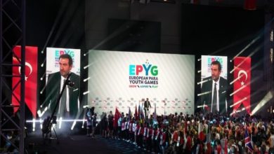 Geniş ve kalabalık bir salondayız. Sahnenin ortasında büyük bir ekran yer alıyor. Ekranda "EPYG – European Para Youth Games (Avrupa Paralimpik Gençlik Oyunları)" yazısı büyük harflerle yer almakta. Ekranın sağ ve sol taraflarında, konuşma yapan Türkiye Milli Paralimpik Komitesi Başkanı Murat Aksu, büyük yakın plan görüntüsü var. Her iki ekranın arkasında kırmızı zemin üzerine beyaz ay-yıldız bulunan Türk bayrağı duruyor. Onun önünde, sahnenin alt kısmında farklı ülkelerden gelen genç sporcular yan yana dizilmiş. Her bir sporcunun elinde ülkelerinin bayrakları veya ülke isimlerinin yazılı olduğu tabelalar var. Katılımcılar arasında tekerlekli sandalyede olan sporcular da bulunuyor. Sahne ışıkları güçlü ve renkli; salonun atmosferi coşkulu ve tören havasında.