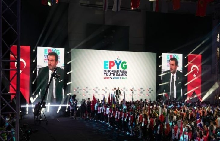 Geniş ve kalabalık bir salondayız. Sahnenin ortasında büyük bir ekran yer alıyor. Ekranda "EPYG – European Para Youth Games (Avrupa Paralimpik Gençlik Oyunları)" yazısı büyük harflerle yer almakta. Ekranın sağ ve sol taraflarında, konuşma yapan Türkiye Milli Paralimpik Komitesi Başkanı Murat Aksu, büyük yakın plan görüntüsü var. Her iki ekranın arkasında kırmızı zemin üzerine beyaz ay-yıldız bulunan Türk bayrağı duruyor. Onun önünde, sahnenin alt kısmında farklı ülkelerden gelen genç sporcular yan yana dizilmiş. Her bir sporcunun elinde ülkelerinin bayrakları veya ülke isimlerinin yazılı olduğu tabelalar var. Katılımcılar arasında tekerlekli sandalyede olan sporcular da bulunuyor. Sahne ışıkları güçlü ve renkli; salonun atmosferi coşkulu ve tören havasında.