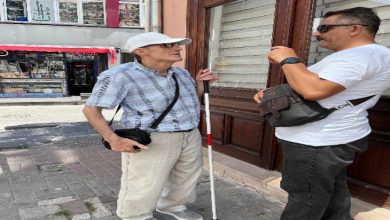 Görselde, bir kaldırım kenarında iki erkek sohbet ederken görülüyor. Soldaki kişi görme engelli; elinde beyaz bastonu var, şapka takıyor ve çapraz askılı bir çanta taşıyor. Sağdaki kişi ise beyaz tişörtlü, omzunda küçük bir çanta var ve karşısındakine dikkatle bir şeyler anlatıyor gibi görünüyor. Arka planda bir dükkân ve çeşitli afişler dikkat çekiyor.