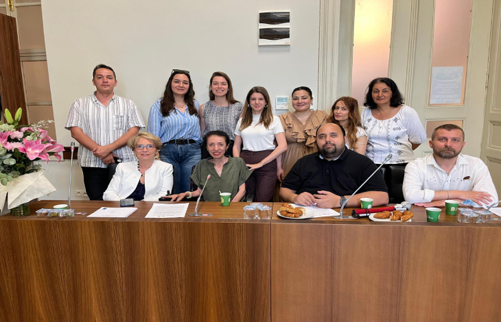 Görselde, bir toplantı veya panel ortamında çekilmiş grup fotoğrafı yer alıyor. Ön sırada masa arkasında oturan 5 kişi bulunuyor, arkalarında ise ayakta duran 8 kişi var. Masada belgeler, mikrofonlar, çay-kahve bardakları ve ikramlıklar (poğaça, kruvasan vb.) dikkat çekiyor. Sol köşede büyük bir çiçek buketi yer almakta. Mekân, yüksek tavanlı ve klasik tarzda bir iç mekân izlenimi veriyor.