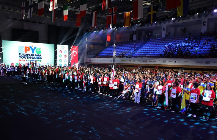 Görselde büyük bir spor salonunda düzenlenen uluslararası bir açılış töreni görülüyor. Sahnenin solunda dev ekranda “EPYG European Para Youth Games” logosu yer alıyor. Alanda farklı ülkelerden gelen sporcular ellerinde ülke bayrakları ve pankartlarla sıraya dizilmiş. Üst kısımda çeşitli ülke bayrakları salonu süslüyor. Katılımcılar arasında tekerlekli sandalye kullanan sporcular da var. Bu sahne, 2025 Avrupa Para Gençlik Oyunları’nın coşkulu açılış anını yansıtıyor.
