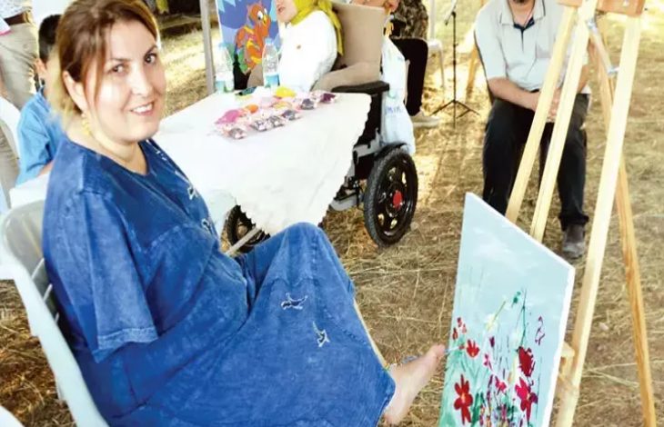 Görselde iki kolu ampute olan orta yaşlarda bir kadın ayak parmağıyla tuttuğu fırçayla resim yapıyor.