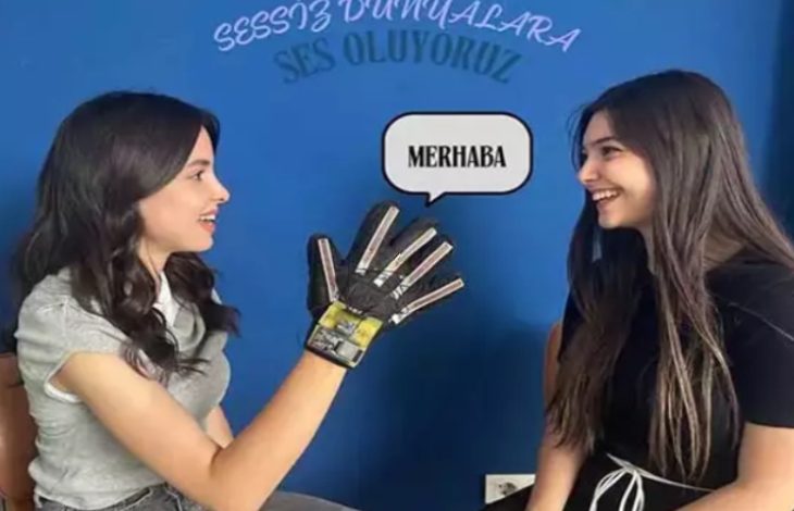 Görselde iki genç kadın birbirine bakarak gülümsüyor. Soldaki kadın eline takılı elektronik sensörlü bir eldivenle işaret dili yapıyor, eldivenden çıkan konuşma balonunda “MERHABA” yazıyor. Arkada mavi duvarda “Sessiz Dünyalara Ses Oluyoruz” ifadesi yer alıyor.