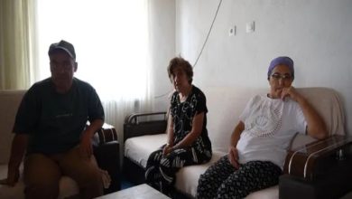Görselde mütevazı bir evin oturma odasında oturan üç kişi görülüyor. Solda ve ortada iki engelli birey, sağda ise başörtülü bir kadın yer almakta. Üçü de sade kıyafetler giymiş, yüz ifadeleri yorgun ama huzurlu. Oda sade döşenmiş; duvarda prizler ve kablolar dikkat çekiyor. Pencereden süzülen doğal ışık mekâna aydınlık katıyor.