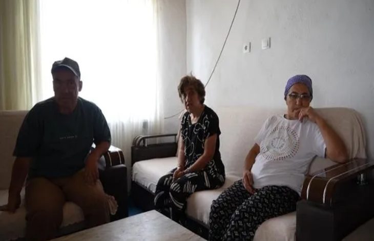 Görselde mütevazı bir evin oturma odasında oturan üç kişi görülüyor. Solda ve ortada iki engelli birey, sağda ise başörtülü bir kadın yer almakta. Üçü de sade kıyafetler giymiş, yüz ifadeleri yorgun ama huzurlu. Oda sade döşenmiş; duvarda prizler ve kablolar dikkat çekiyor. Pencereden süzülen doğal ışık mekâna aydınlık katıyor.