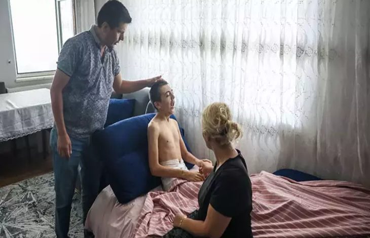 Görselde, ev ortamında bir yatağın üzerinde oturan genç bir erkek, yanında oturan bir kadın ve ayakta duran bir adamla birlikte görülüyor. Kadın gencin ellerini tutarken, adam ise gencin başına dokunuyor. Oda, gündüz ışığı alan beyaz tül perdeli bir pencereden aydınlanıyor.