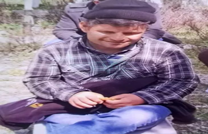 Fotoğrafta, açık havada çekilmiş bir karede, kareli gömlek, mavi kot pantolon ve siyah bere takan bir Tunay Çetiner oturuyor. Kucağında koyu renk bir çanta bulunuyor ve ellerinde küçük bir nesne tutuyor. Arka planda bulanık şekilde başka insanlar ve doğa manzarası görünüyor.