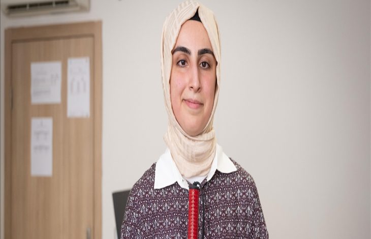 Görselde, başörtülü genç bir kadın Ayşe Begüm Başbozkurt Gür iç mekânda kameraya doğru gülümseyerek poz veriyor. Üzerinde desenli bir tunik ve beyaz yakalı bir gömlek bulunuyor. Boynunda bir mikrofon ya da ses cihazı klipsi var, bu da bir röportaj ya da tanıtım çekimi olabileceğini düşündürüyor. Arka planda kapalı bir kapı ve duvarda bazı kağıtlar asılı.
