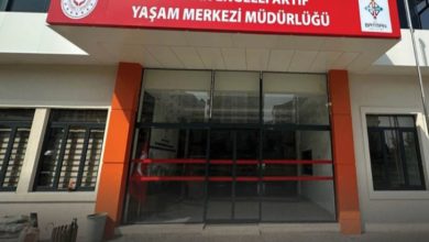 Görselde, **Batman Engelli Aktif Yaşam Merkezi Müdürlüğü** binasının ana girişi görülüyor. Üstte kırmızı zemin üzerine beyaz harflerle yazılmış kurum adı yer alıyor. Giriş kapısı camdan ve otomatik; yanlarda turuncu-siyah sütunlar bulunuyor. Sol üst köşede Aile ve Sosyal Hizmetler Bakanlığı logosu, sağ üst köşede ise Batman Belediyesi logosu yer almakta. Yapı modern ve düzenli bir mimariye sahip.