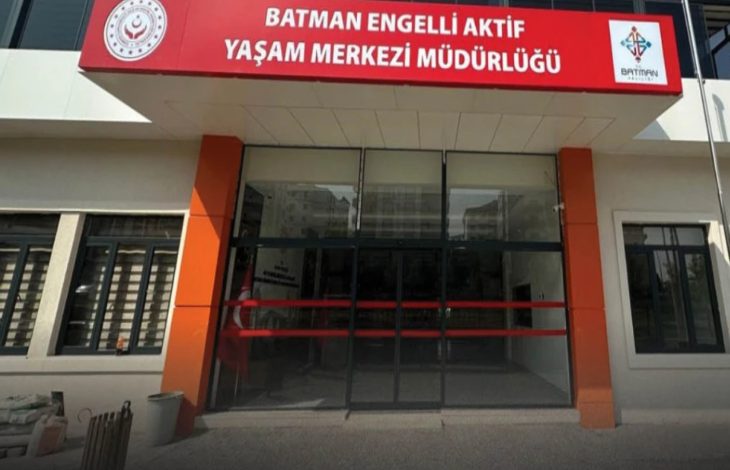Görselde, **Batman Engelli Aktif Yaşam Merkezi Müdürlüğü** binasının ana girişi görülüyor. Üstte kırmızı zemin üzerine beyaz harflerle yazılmış kurum adı yer alıyor. Giriş kapısı camdan ve otomatik; yanlarda turuncu-siyah sütunlar bulunuyor. Sol üst köşede Aile ve Sosyal Hizmetler Bakanlığı logosu, sağ üst köşede ise Batman Belediyesi logosu yer almakta. Yapı modern ve düzenli bir mimariye sahip.