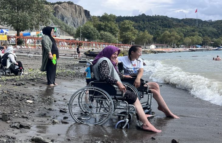 Bolu Belediyesi’nin düzenlediği etkinlik sayesinde hayatında ilk kez denizi gören 76 yaşındaki Huriye Çelik, sahilde mutluluğun tadını çıkarıyor. Bir sahil kenarında çekilmiş bu fotoğrafta, tekerlekli sandalyedeki yaşlı bir kadın ve yanındaki genç kadın birlikte deniz kıyısında oturuyor. Kadının ayağına dalgalar çarpıyor, yüzünde ise neşe dolu bir gülümseme var. Arka planda başka tekerlekli sandalye kullanıcıları ve onları izleyen insanlar görülüyor.
