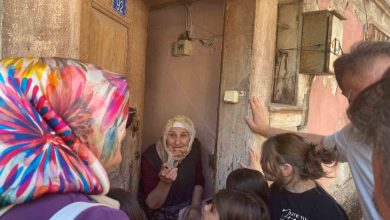 Görselde, başörtülü yaşlı bir kadın, kapısının önünde elini uzatarak konuşuyor. Etrafında başörtülü bir kadın, birkaç çocuk ve bir erkek bulunuyor. Kapının üzerinde “92” numarası yer alıyor, bina ise eski ve yıpranmış görünüyor. Ortam, mahallede yapılan samimi bir ziyaret havasında.