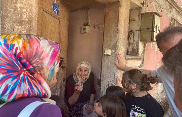 Görselde, başörtülü yaşlı bir kadın, kapısının önünde elini uzatarak konuşuyor. Etrafında başörtülü bir kadın, birkaç çocuk ve bir erkek bulunuyor. Kapının üzerinde “92” numarası yer alıyor, bina ise eski ve yıpranmış görünüyor. Ortam, mahallede yapılan samimi bir ziyaret havasında.