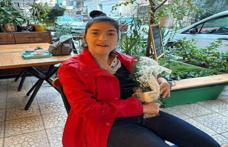 Görselde, genç bir kadın bir kafede oturuyor. Üzerinde kırmızı bir mont, koyu renk pantolon ve siyah üst var. Elinde beyaz çiçeklerden oluşan bir buket tutuyor. Arka planda yeşil bitkiler, ahşap masa-sandalyeler ve camın dışından görünen park halindeki bir araç yer alıyor.