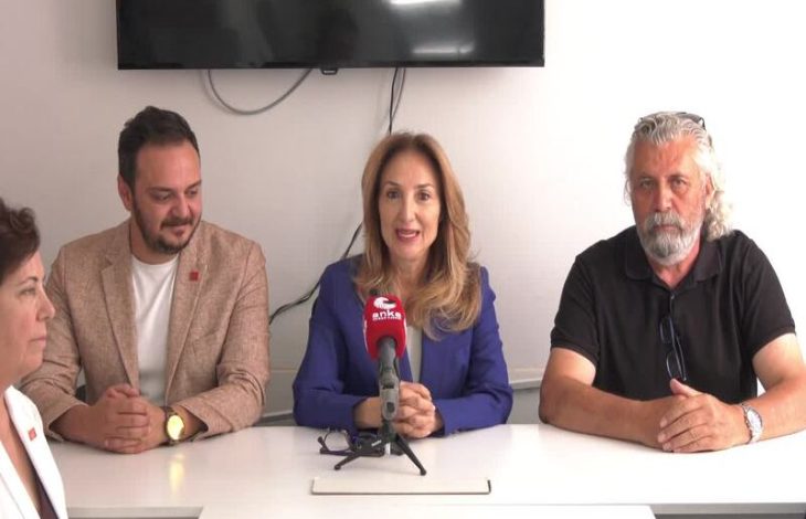 Görselde bir basın toplantısı görüntüsü yer alıyor. Ortada mavi ceketli Aylin Nazlıaka mikrofon önünde konuşuyor. Yanında üç kişi daha bulunuyor; solda açık renk ceketli bir erkek ve beyaz ceketli bir kadın, sağda ise siyah tişörtlü beyaz saçlı bir erkek oturuyor. Masanın üzerinde mikrofon var, arka planda ise duvara asılı bir televizyon ekranı görülüyor.