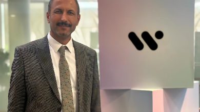 Görselde, WeAccess.AI CEO’su Doç. Dr. Özer Çelik, firmanın logosunun önünde takım elbisesi ile birlikte poz veriyor.