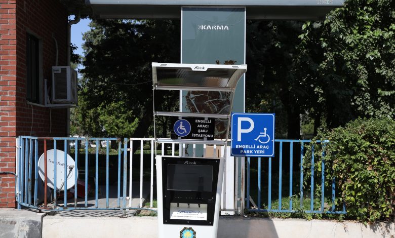 Bir bina önünde, Diyarbakır Büyükşehir Belediyesi'ne ait "Engelli Araç Şarj İstasyonu" ve "Engelli Araç Park Yeri" tabelalarının bulunduğu bir kiosk görülüyor. Kioskun üstünde güneşlik, arkasında ise ağaçlar ve bir çit yer alıyor.